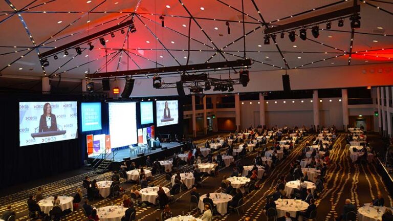 A large conference hall filled with attendees sits under a geometric ceiling with red lighting