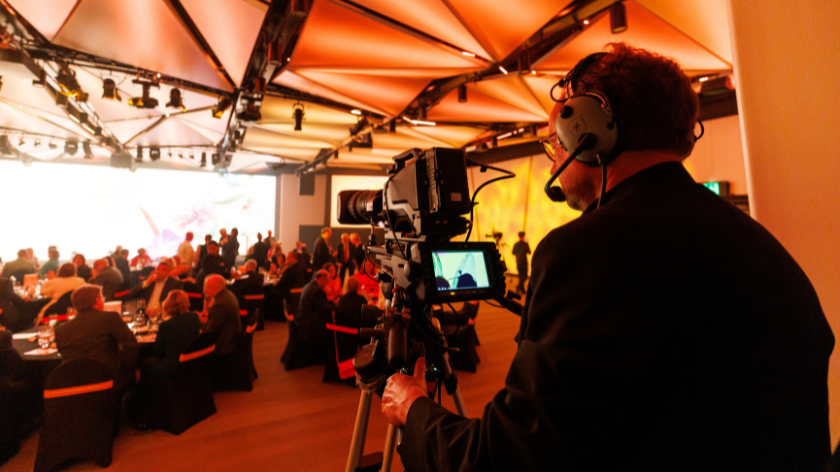 Image features an AV team member operating a camera filming a gala dinner and speakers presenting on stage.