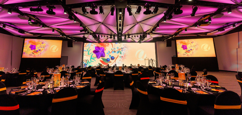 Boulevard room with banquet tables set for a gala dinner. The ceiling shows pink, mosaic style feature lighting. The front of the room displays a large screen in the center with two smaller screens either side mirroring the middle screen.