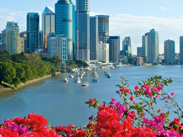 A vibrant panorama of Brisbane city unfolds across a river dotted with sailboats, framed by lush greenery and colorful flowers.