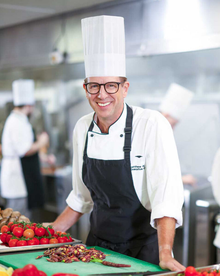 Executive Chef Matthew Arnold beams with a white toque, glasses, and a black apron over his chef's coat.