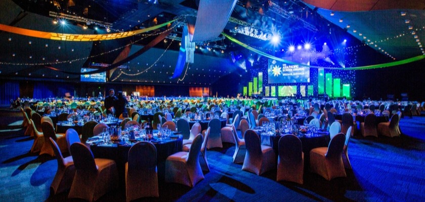 The Great Hall hosting over 50 banquet tables set for dinner. The ceiling features string lights and long fabric strips connecting in the center of the room, the front of the room displays a large screen.