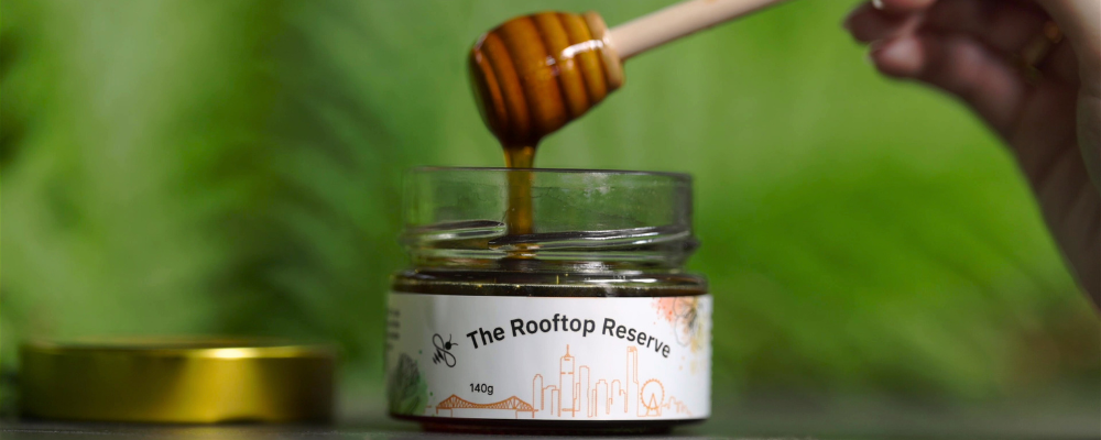 Image of a jar of honey with a honey wand dipping into the jar. The label displays The Rooftop Reserve text with an outline of the Brisbane city skyline.
