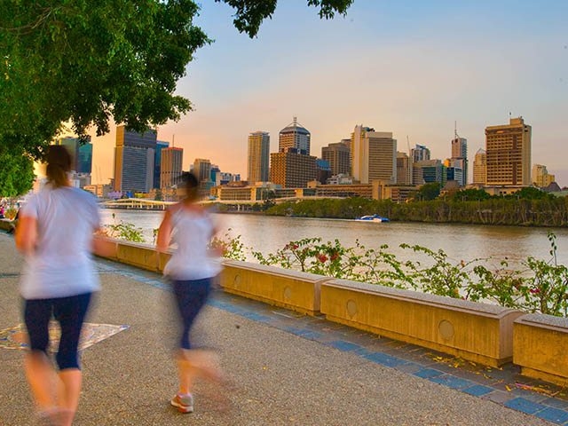 Two people jog along a riverfront path in Brisbane, Queensland, with the city skyline in the background.